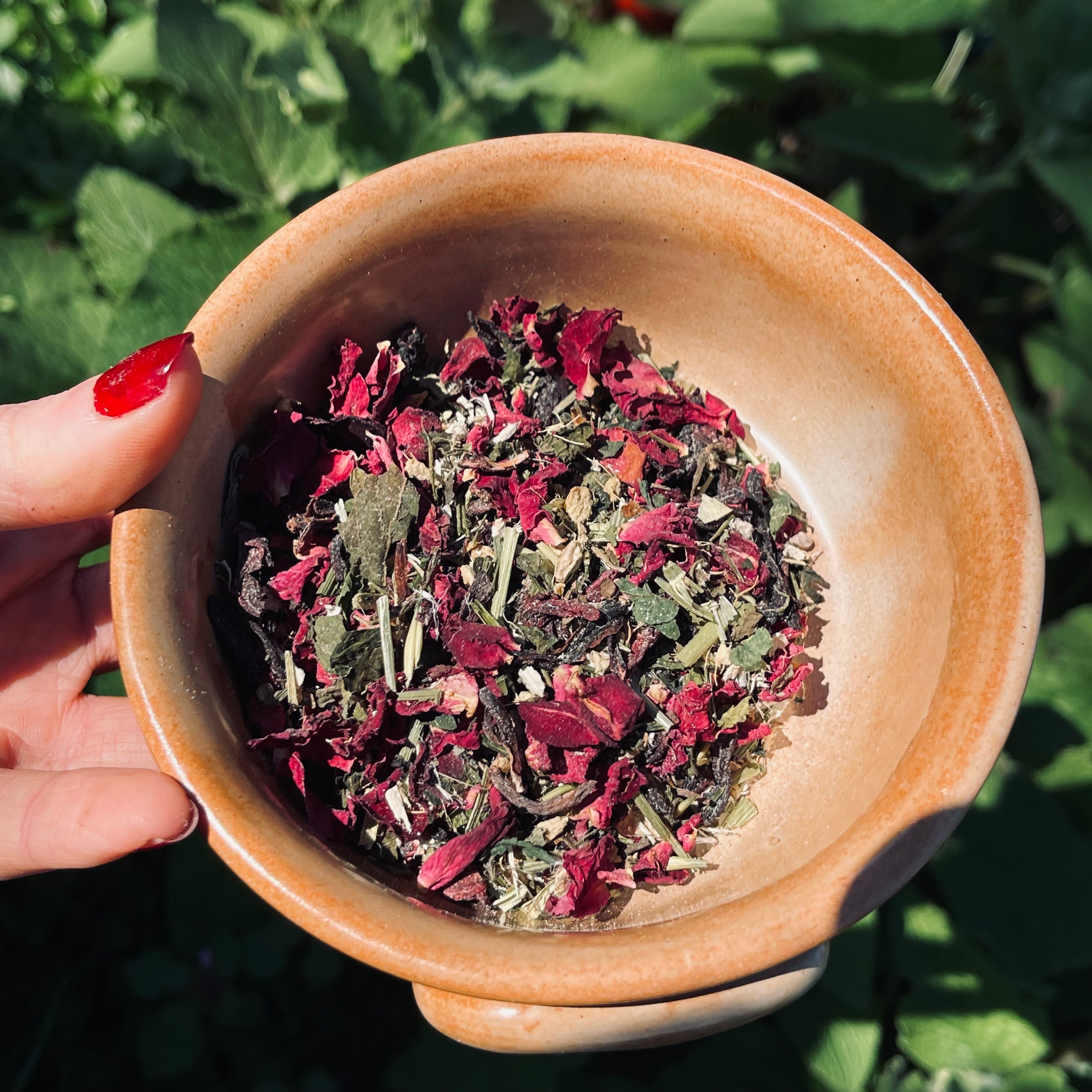 Bowl of dried herbs held by a hand with red nail polish against a green leafy background